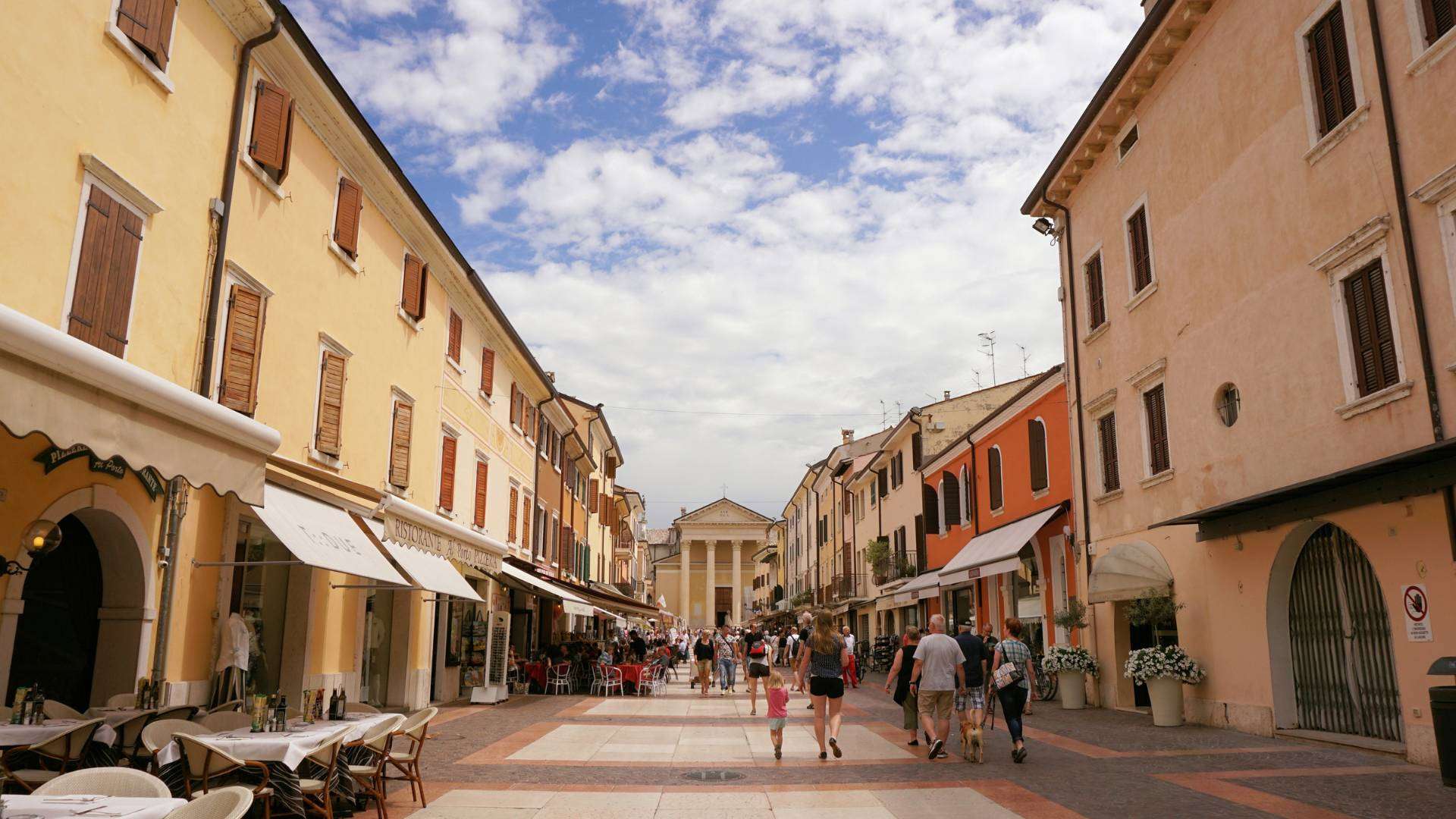 Belebte Fußgängergasse in Bardolino mit Cafés, Arkaden und Kirchenfassade am Ende