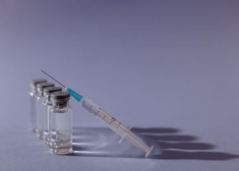 Spritze liegt neben mehreren Impfampullen auf grauem Untergrund