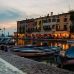 Hölzerne Fischerboote im Hafen von Lazise, dahinter beleuchtete Häuserfassaden am Abend