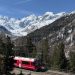 Roter Zug der Rhätischen Bahn fährt durch ein Hochtal vor schneebedeckten Gipfeln des Berninamassivs
