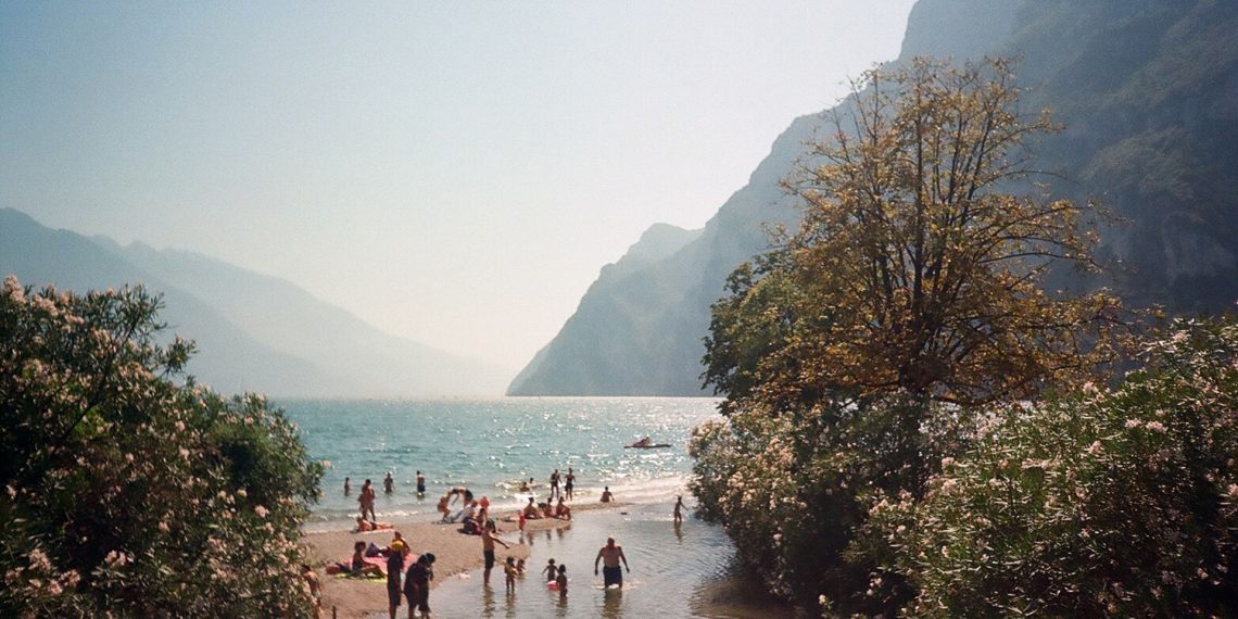 Badegäste schwimmen und sonnen sich an einem Ufer des Gardasees vor der Bergkulisse