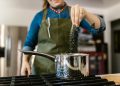 Eine Frau in Schürze streut mit der Hand Salz in einen Kochtopf auf dem Gasherd, ohne abzumessen
