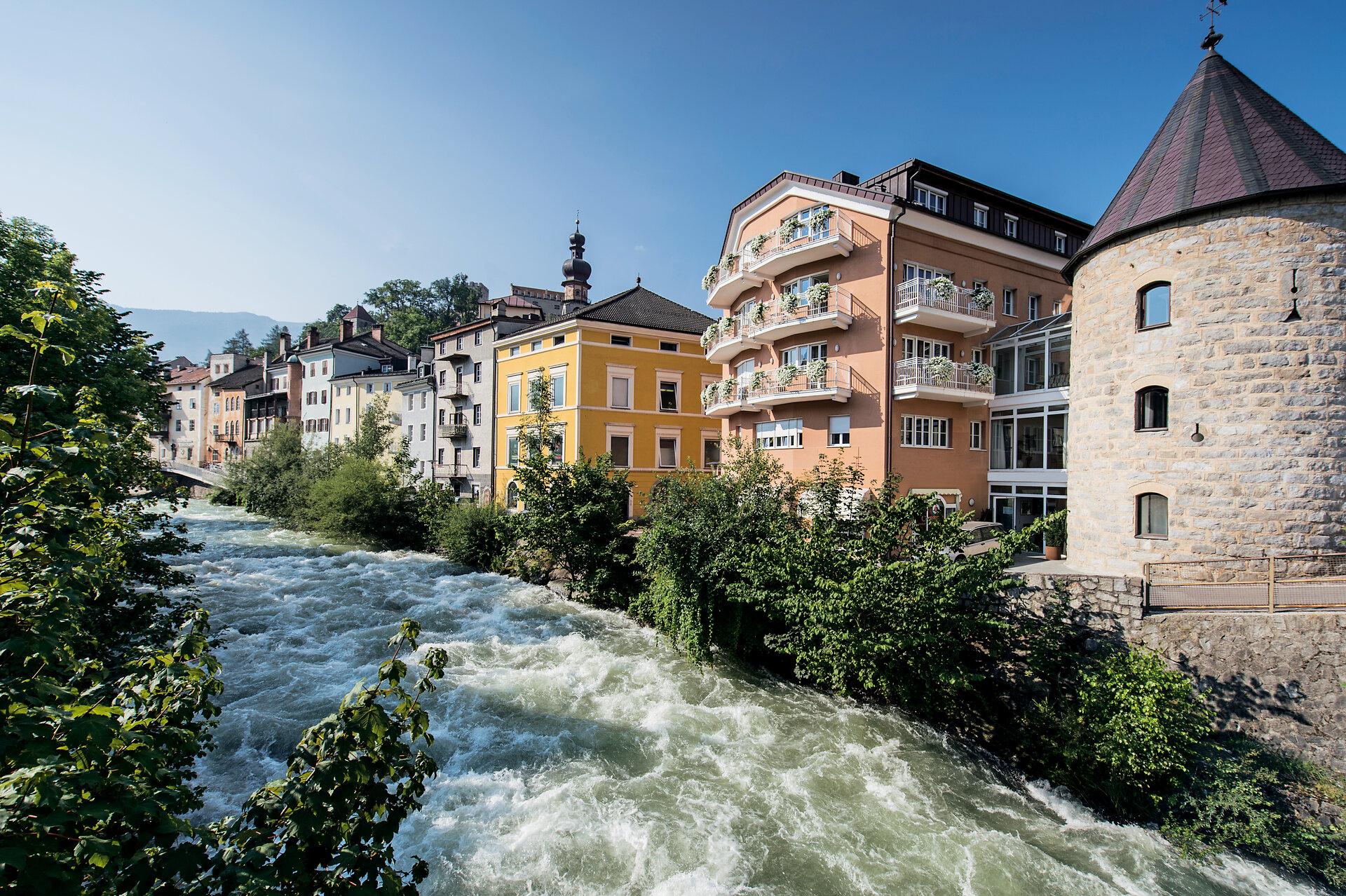Die Rienz fließt an bunten Häusern und einem runden Turm in Bruneck vorbei