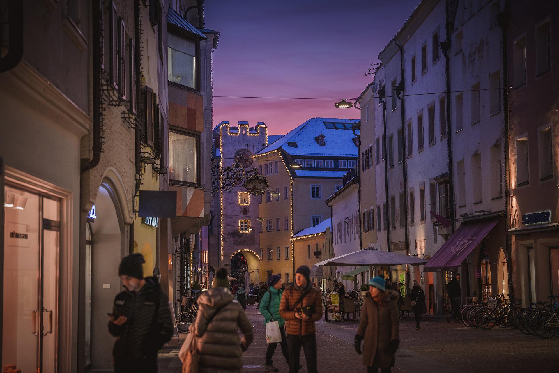 Die Brunecker Stadtgasse in der Abenddämmerung im Winter, mit beleuchteten Schaufenstern und Passanten