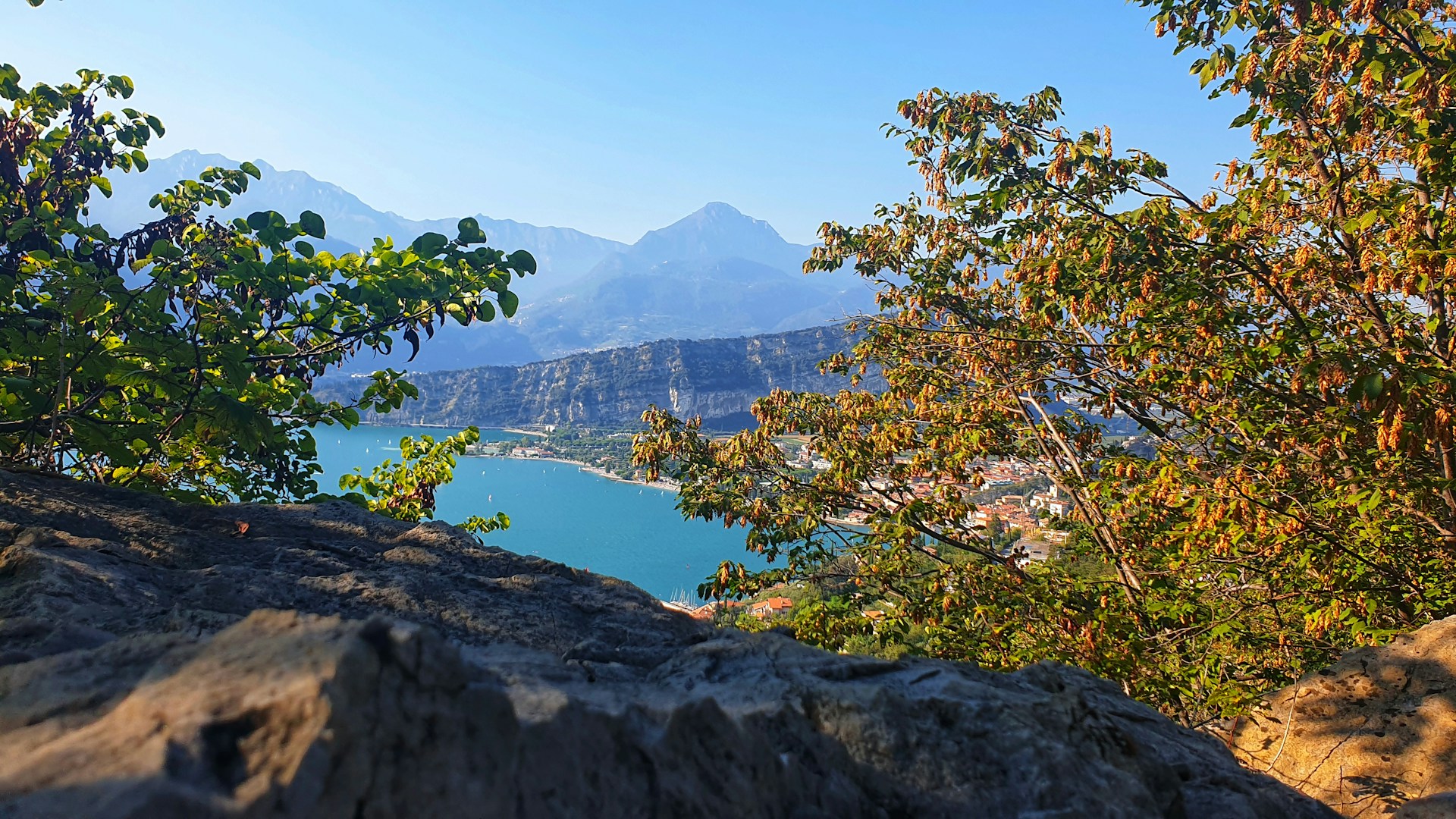 Blick zwischen Bäumen vom Wanderweg Busatte-Tempesta auf den Gardasee und die Berge am Westufer