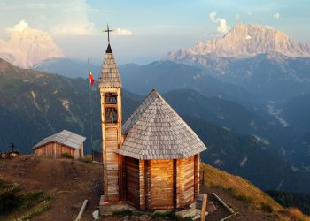 Holzkapelle mit Glockenturm auf dem Gipfel des Col di Lana, dahinter Dolomitengipfel im Abendlicht