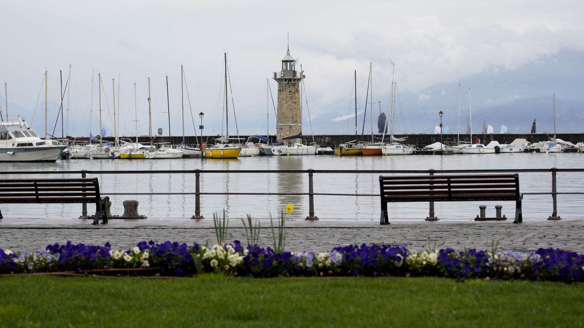Leuchtturm und Segelboote im Hafen von Desenzano del Garda, davor Bänke und Frühlingsblumen
