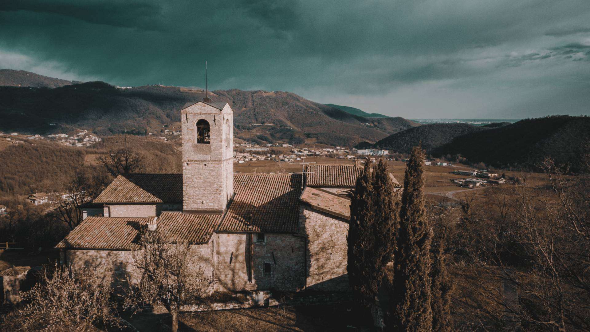 Franciacorta: Romanische Kirche zwischen Zypressen Romanische Kirche mit Glockenturm und Zypressen auf einem Hügel, dahinter Dörfer und Berge unter dunklem Himmel