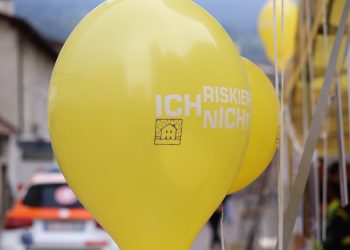 Menschen stehen an Informationsständen und unterhalten sich, im Hintergrund sind Gebäude und Bäume zu sehen. Die Szene zeigt eine Veranstaltung im Freien.