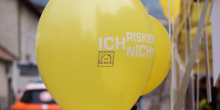 Menschen stehen an Informationsständen und unterhalten sich, im Hintergrund sind Gebäude und Bäume zu sehen. Die Szene zeigt eine Veranstaltung im Freien.