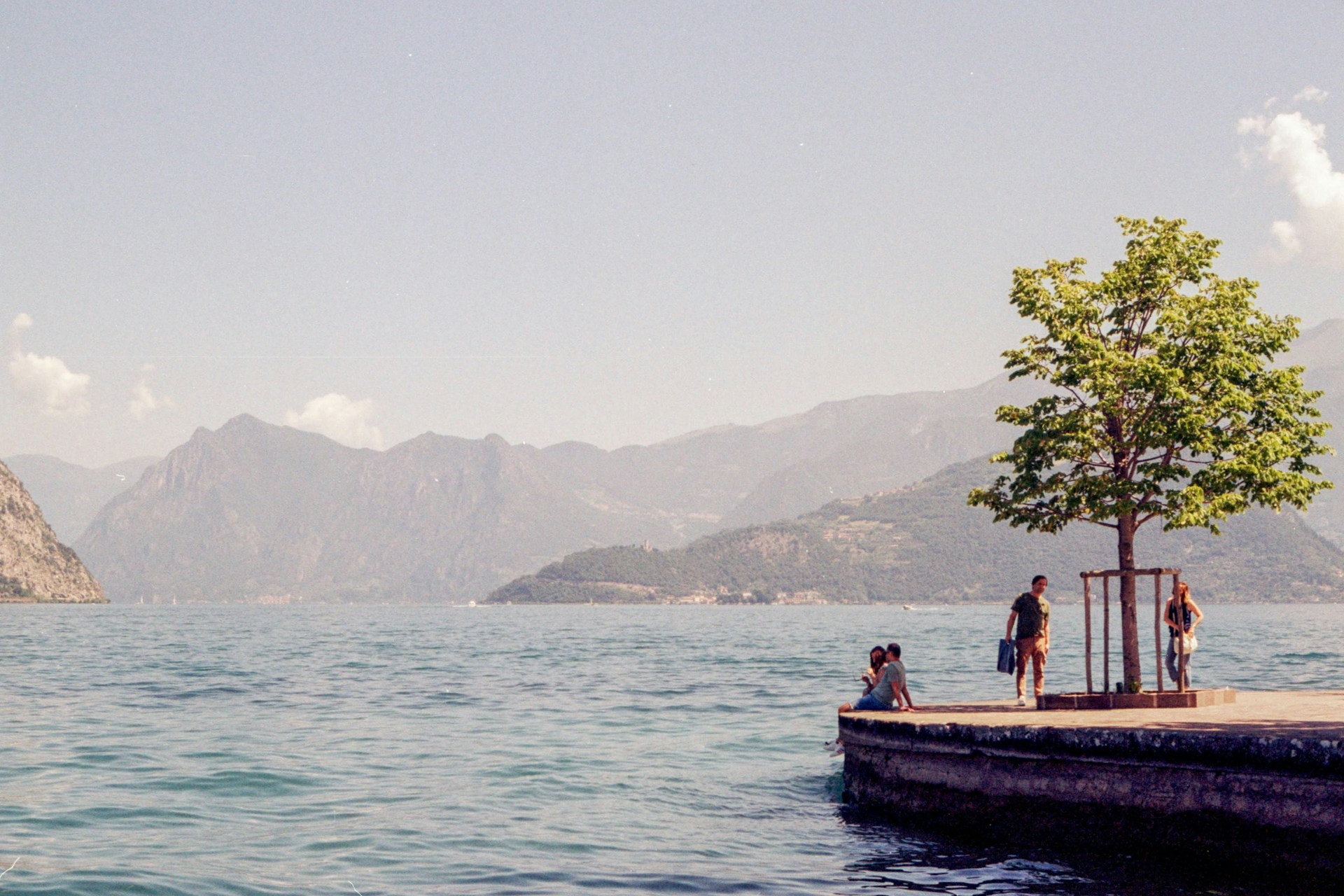 Pause am Wasser am Iseosee Menschen entspannen auf einer Uferplattform mit Baum am Iseosee, dahinter Berge im Dunst