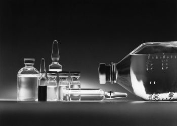 Schwarzweißaufnahme mehrerer Arzneimittelampullen und einer Infusionsflasche auf einer dunklen Fläche