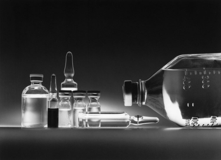 Schwarzweißaufnahme mehrerer Arzneimittelampullen und einer Infusionsflasche auf einer dunklen Fläche