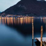 Lichter eines Uferdorfs auf Monte Isola am Abend, im Vordergrund ein Holzsteg im Iseosee