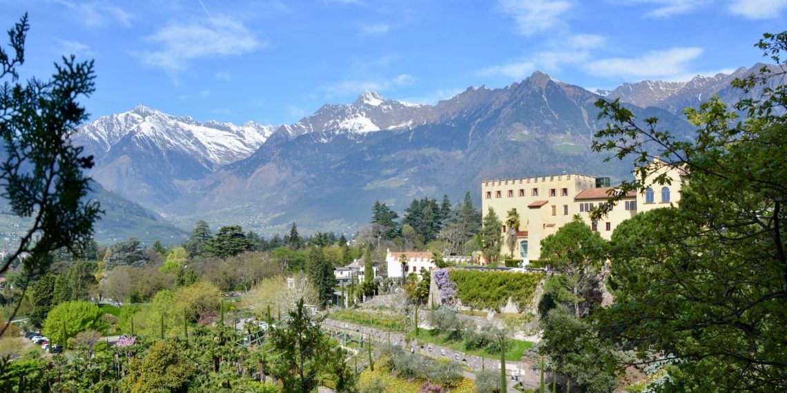 Schloss Trauttmansdorff mit seinen Gärten über Meran, dahinter schneebedeckte Gipfel unter blauem Himmel